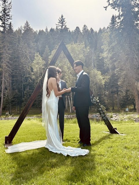 A couple exchanging vows under a timber A-frame arch, backlit by the evening sun