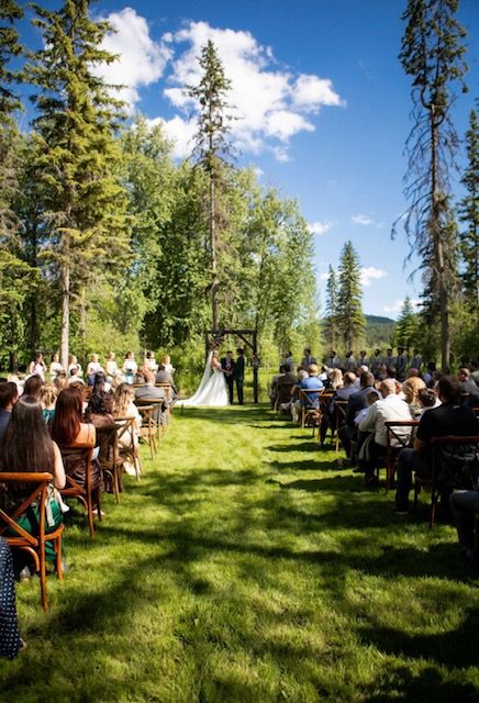 Guests seated in rows on the lawn under tall pines, watching a ceremony at the end of the aisle